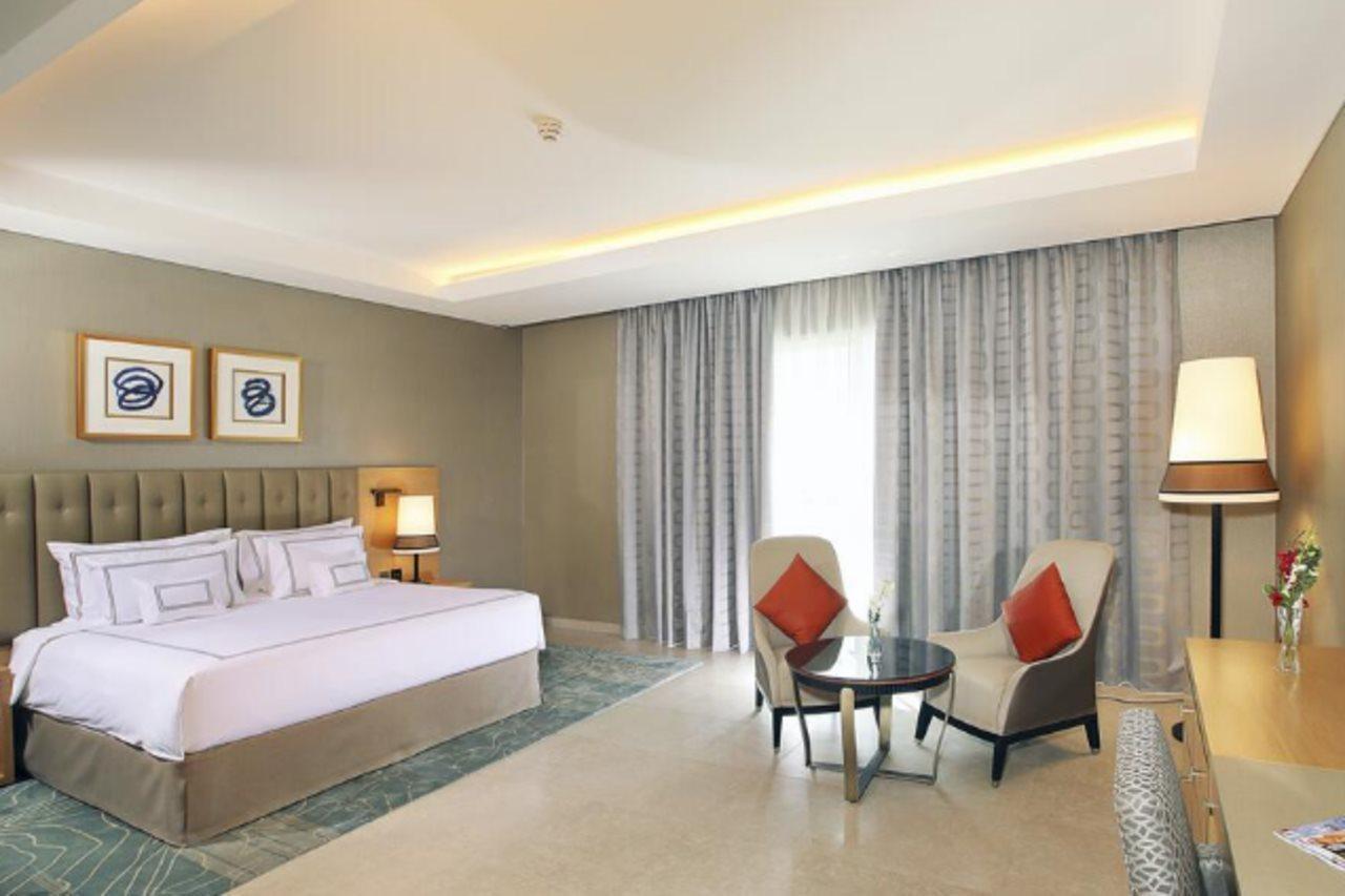 迪拜大都会大酒店 外观 照片 The photo shows a modern hotel room featuring a spacious layout. There is a large bed with a headboard adorned with decorative artwork above it. On one side of the bed, there's a bedside lamp. Opposite the bed, there are two comfortable armchairs sty