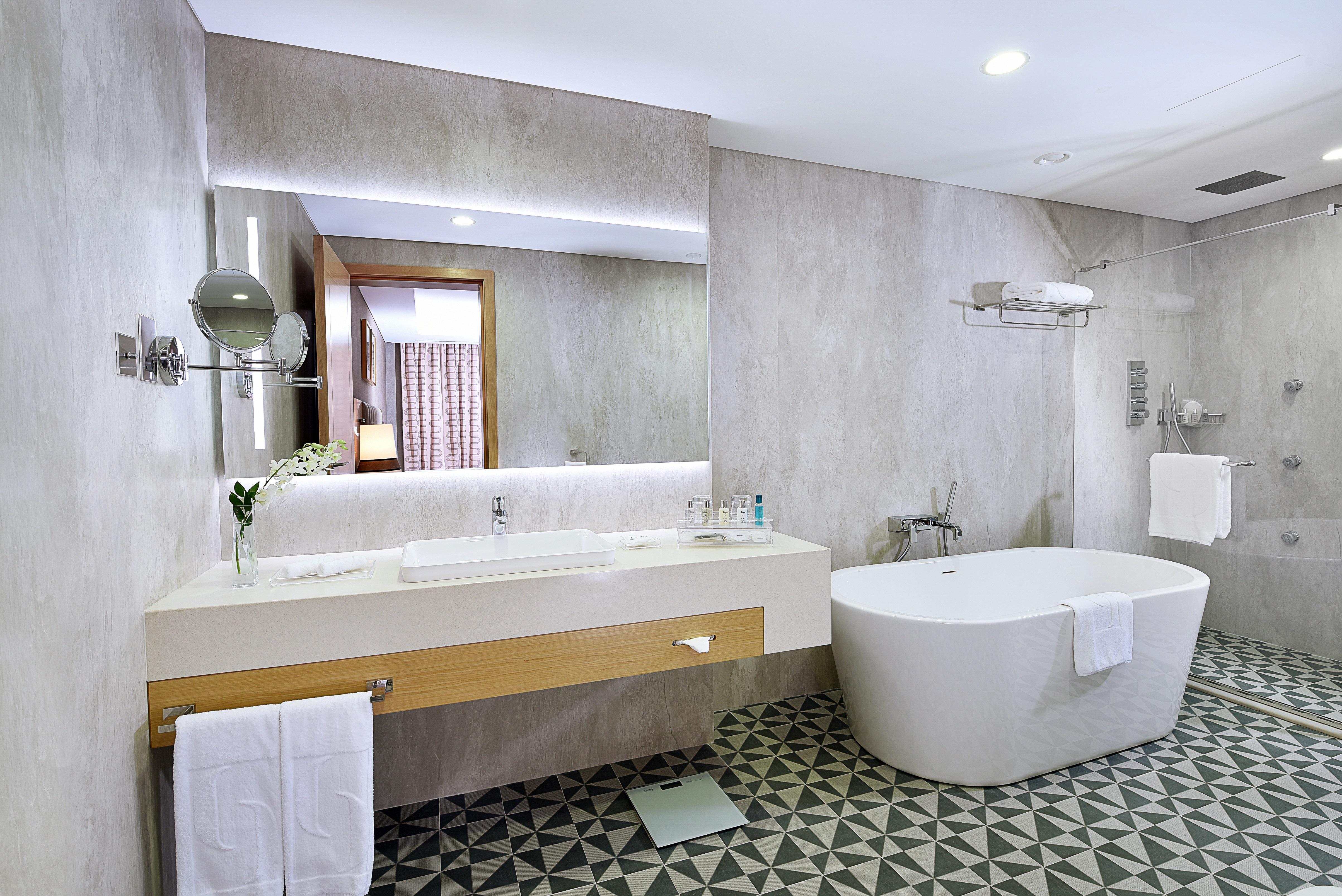 迪拜大都会大酒店 外观 照片 The photo shows a modern bathroom featuring a sleek design. There is a large white freestanding bathtub on one side. The vanity area has a long countertop with a sink and a large mirror above it, illuminated by soft lighting. The walls are a light gr