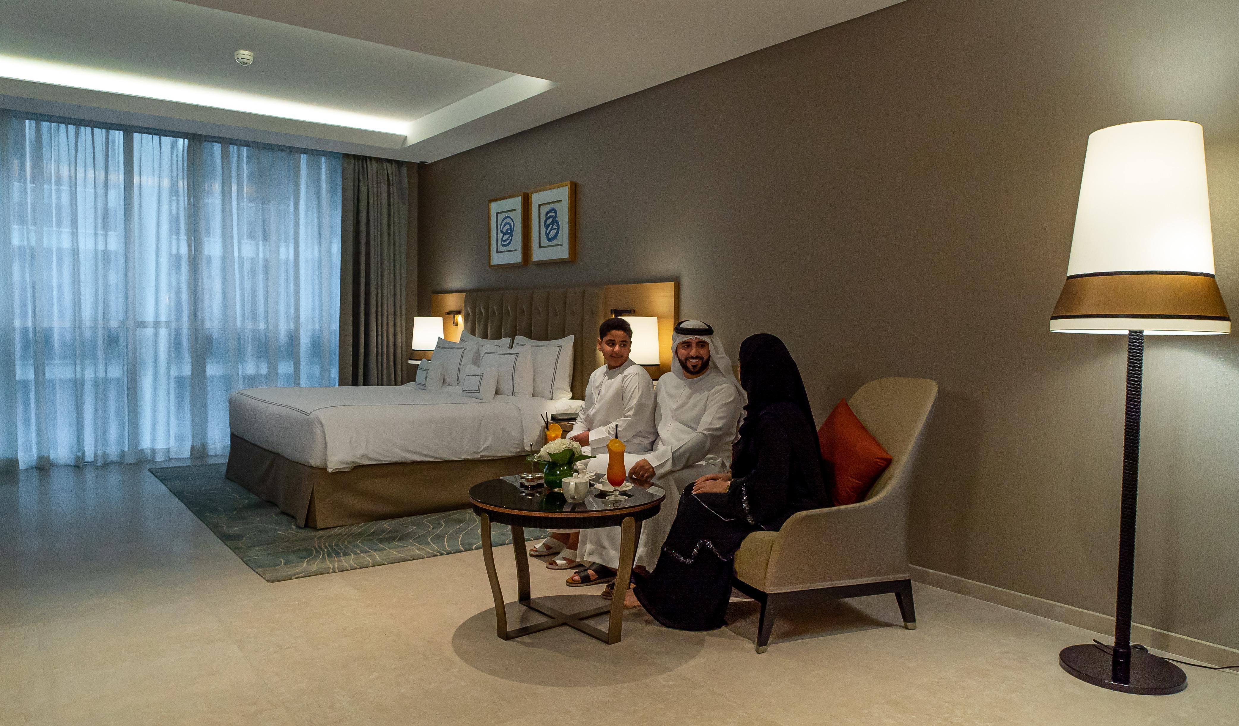 迪拜大都会大酒店 外观 照片 The photo shows a modern hotel room featuring a large bed with white linens and a stylish headboard. In the room, there is a small round table with a floral centerpiece and drinks. Three individuals are seated on the chairs by the table: two adults a