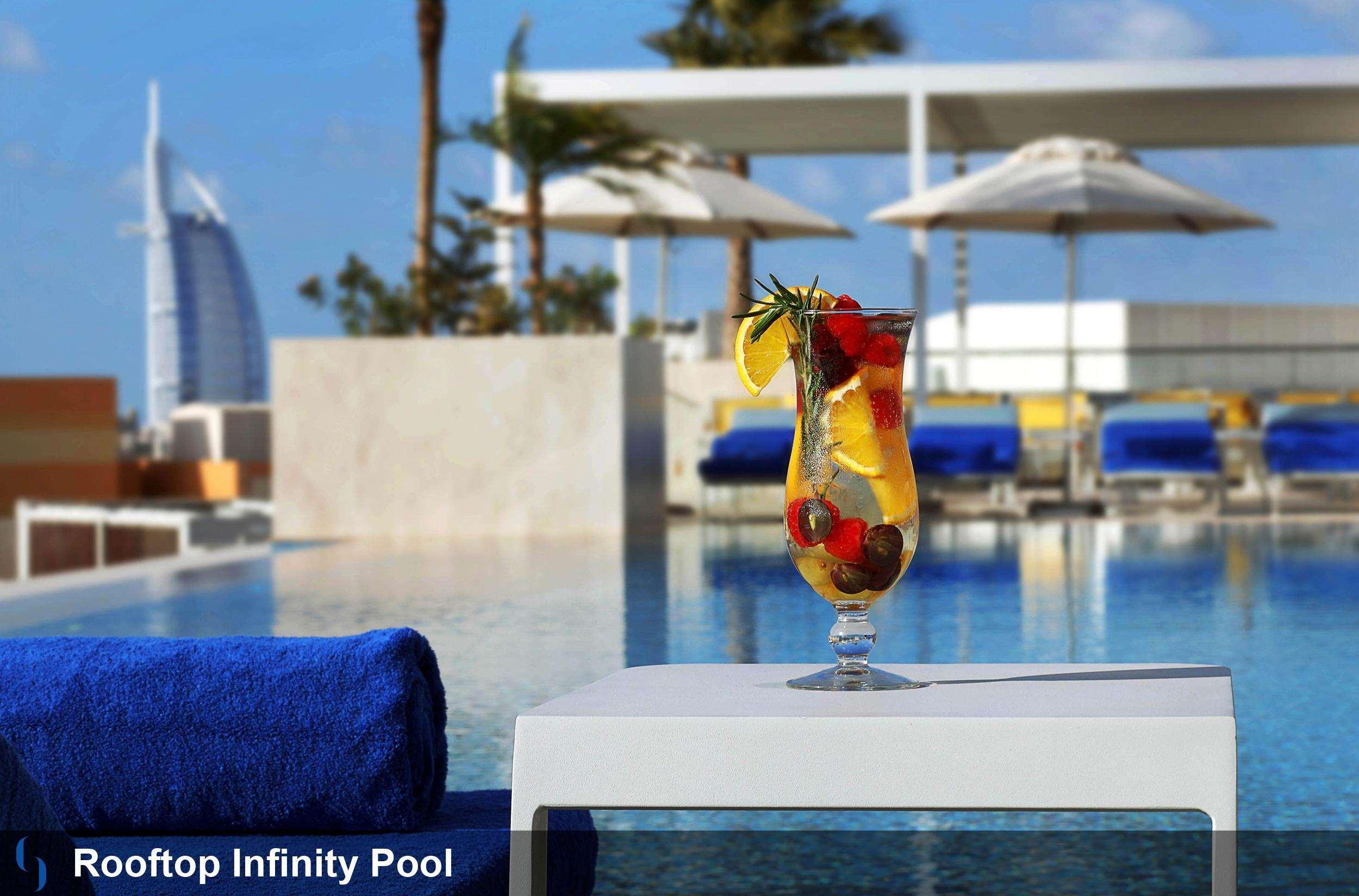迪拜大都会大酒店 外观 照片 The photo shows a rooftop infinity pool with a stunning view. In the foreground, there is a cocktail glass filled with a colorful drink garnished with fruits, including lemon and berries. The background features cozy loungers and umbrellas, suggestin