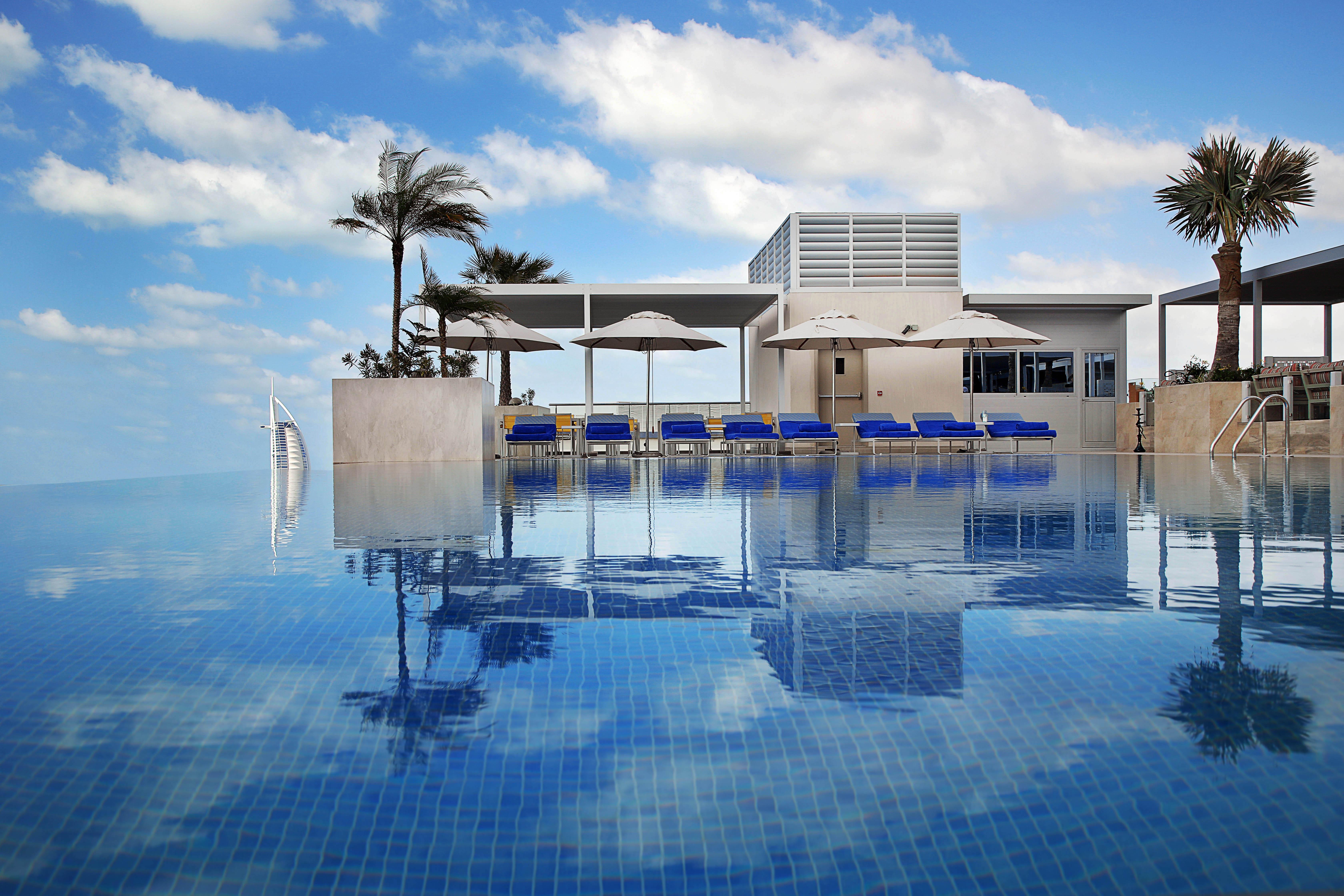 迪拜大都会大酒店 外观 照片 The photo depicts a luxurious swimming pool with a stunning infinity edge. The water is calm and reflects the sky, creating a serene atmosphere. Surrounding the pool are several blue lounge chairs with white umbrellas, providing spots for relaxation.