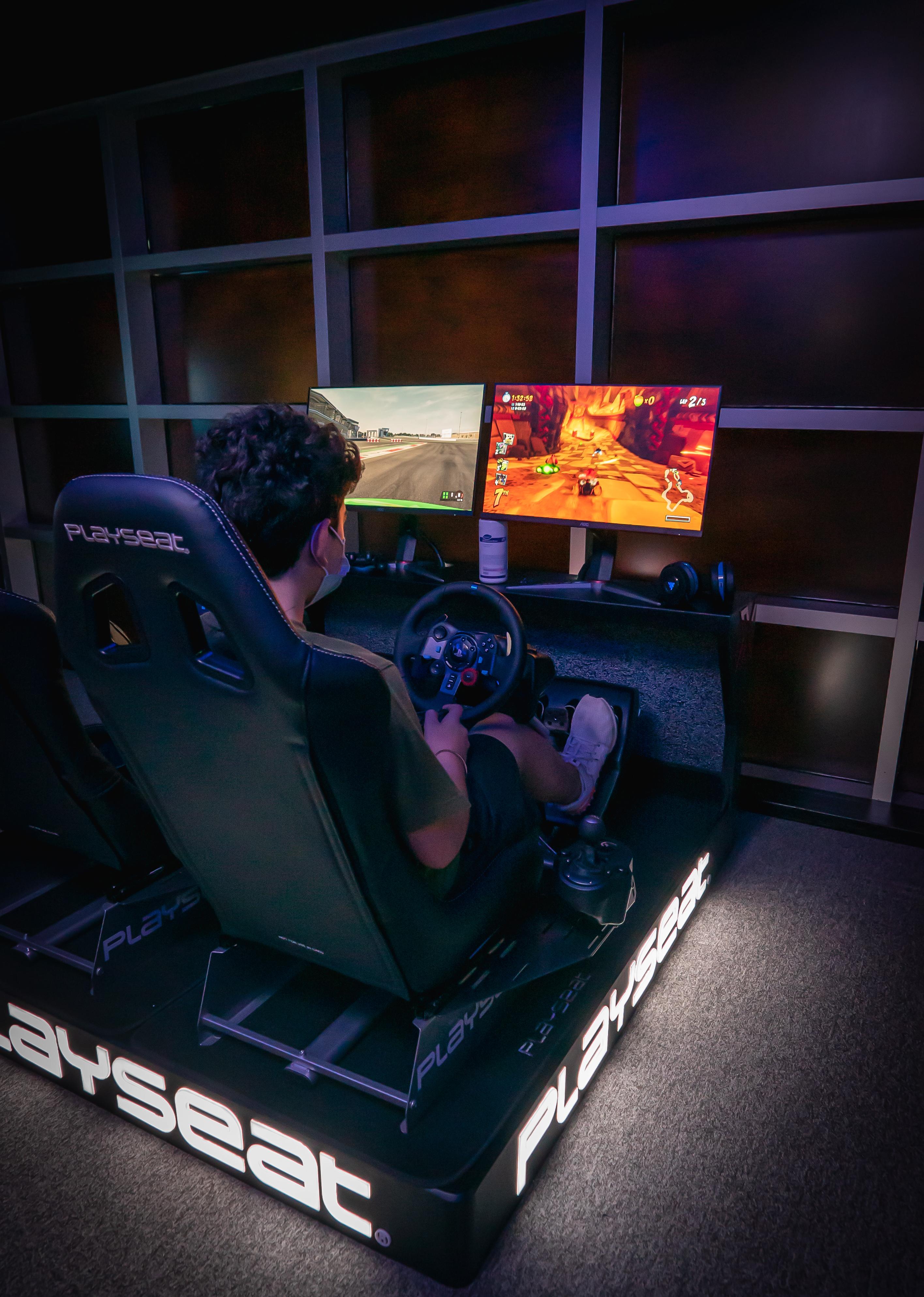 迪拜大都会大酒店 外观 照片 The photo shows a person sitting in a racing gaming chair equipped with a steering wheel and pedals. They are facing two computer monitors. One monitor displays a racing simulation, while the other appears to show a different game or racing scene. Th