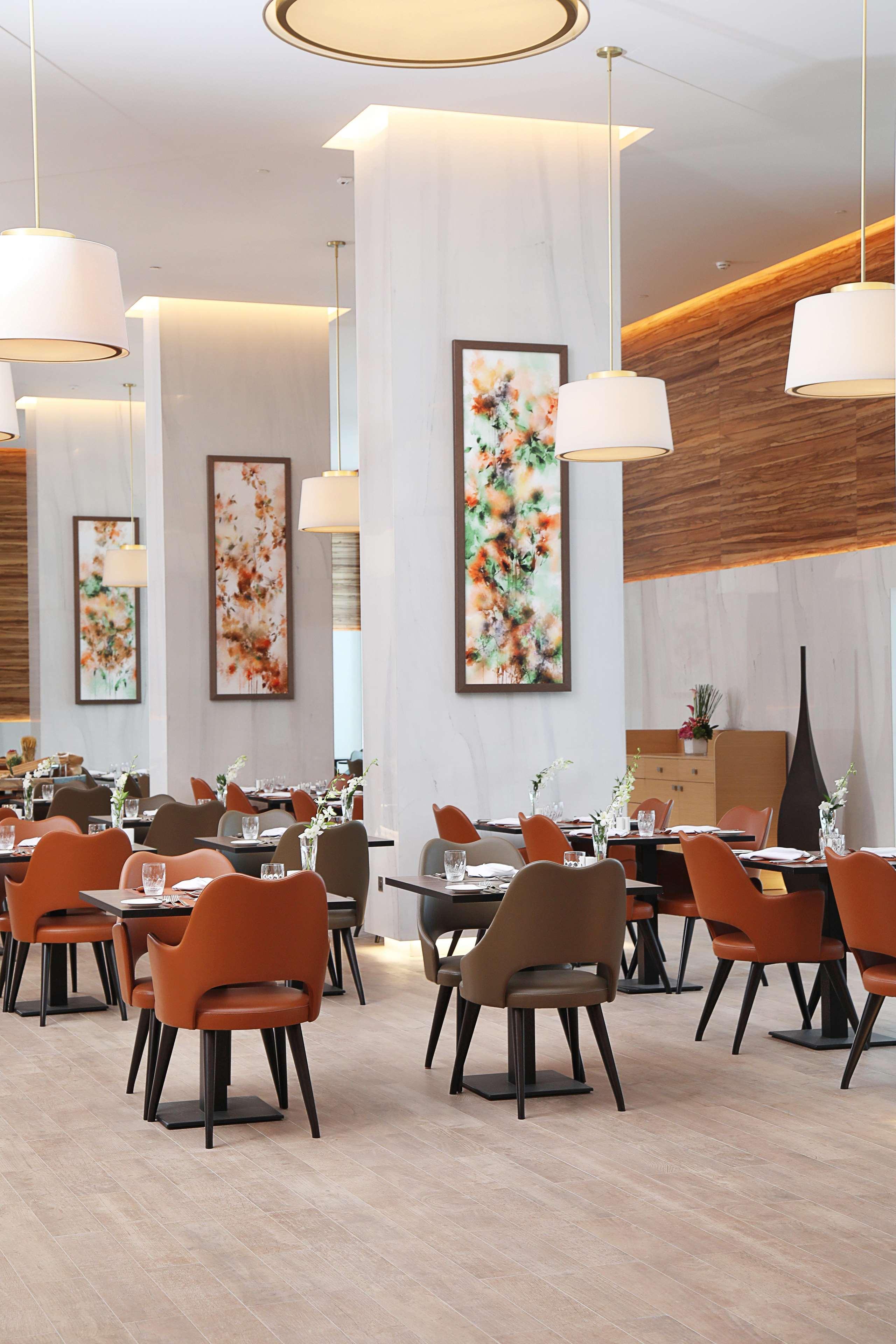 迪拜大都会大酒店 外观 照片 The photo shows the interior of a modern restaurant. The space features elegantly set dining tables with glasses and cutlery, and the chairs have a mix of colors, including orange and neutral tones. There are large pieces of artwork on the walls, lik