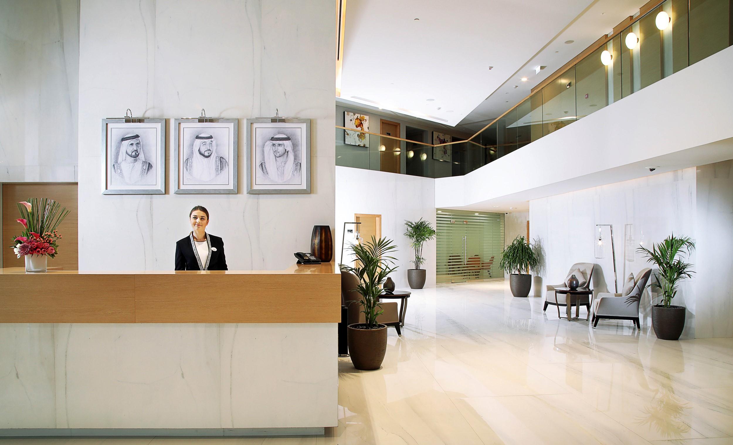 迪拜大都会大酒店 外观 照片 The photo depicts a modern hotel lobby. In the foreground, there is a reception desk with a staff member dressed in formal attire, likely a receptionist. Behind the reception desk, three framed portraits are hanging on the wall, featuring individuals