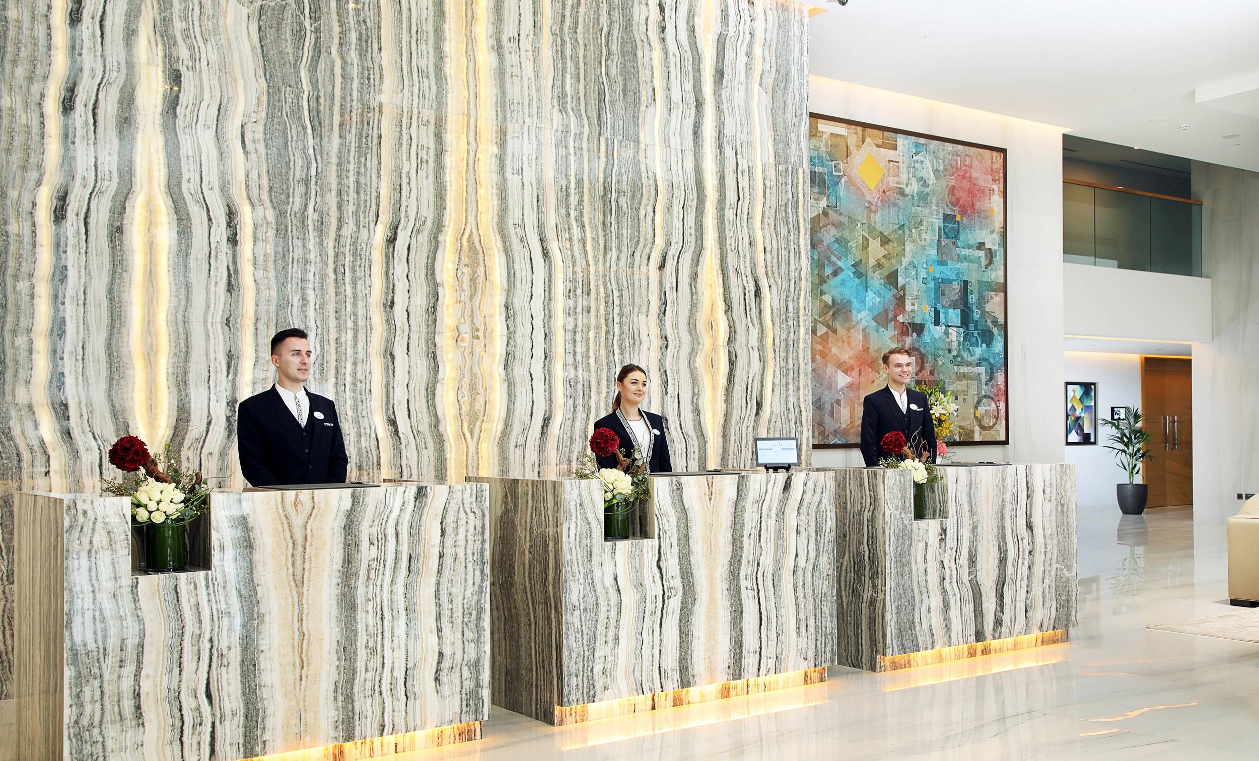 迪拜大都会大酒店 外观 照片 The photo shows a hotel reception area featuring three receptionists standing behind a sleek, modern front desk made of a striped marble-like material with illuminated edges. They are dressed in formal uniforms. Behind them, there is a large piece of