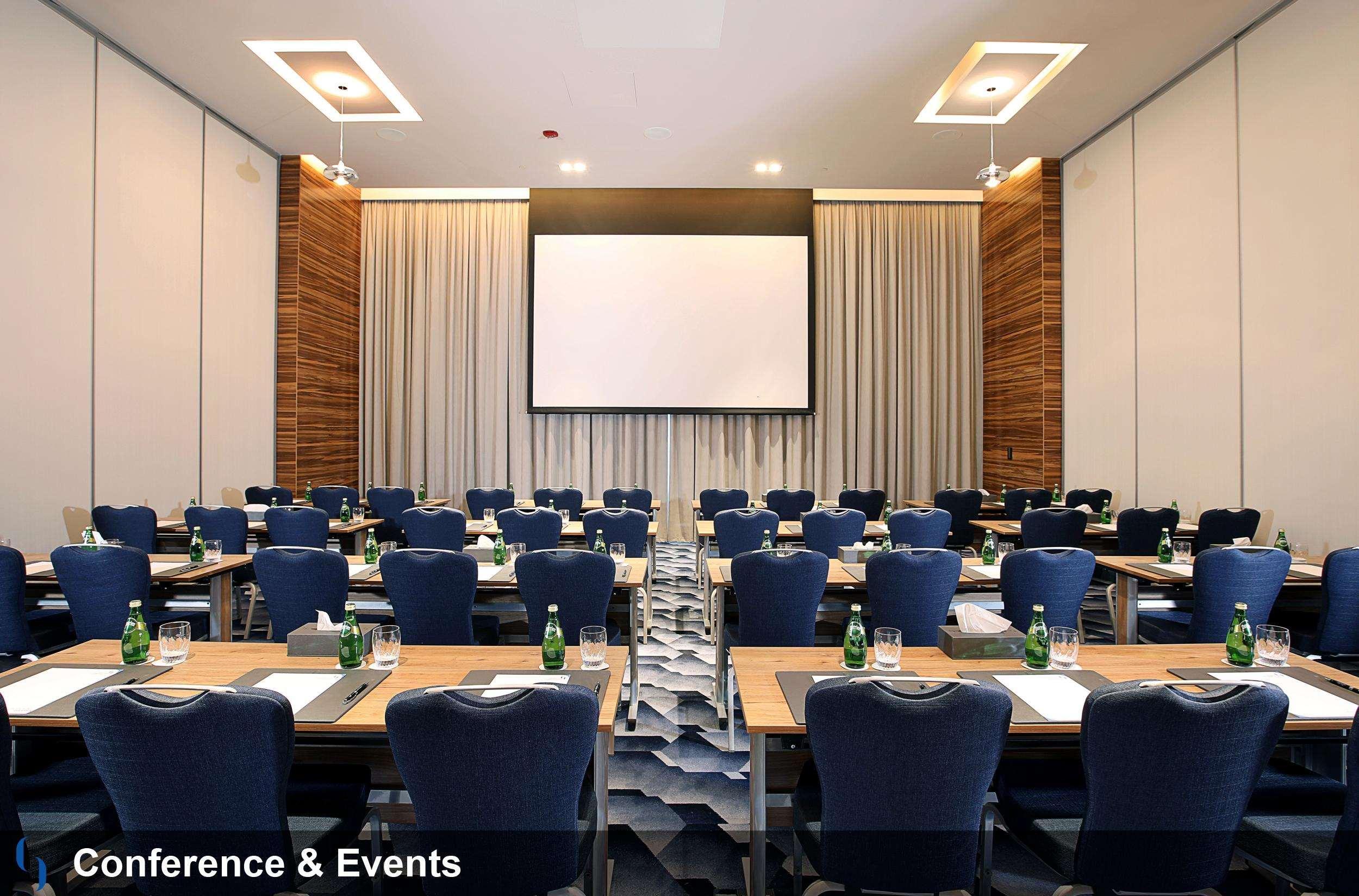 迪拜大都会大酒店 外观 照片 The image shows a conference room set up for an event. The room features rows of tables arranged with blue chairs, and each table has a small bottle of water. At the front, there's a blank projector screen mounted on the wall, flanked by curtains. Th