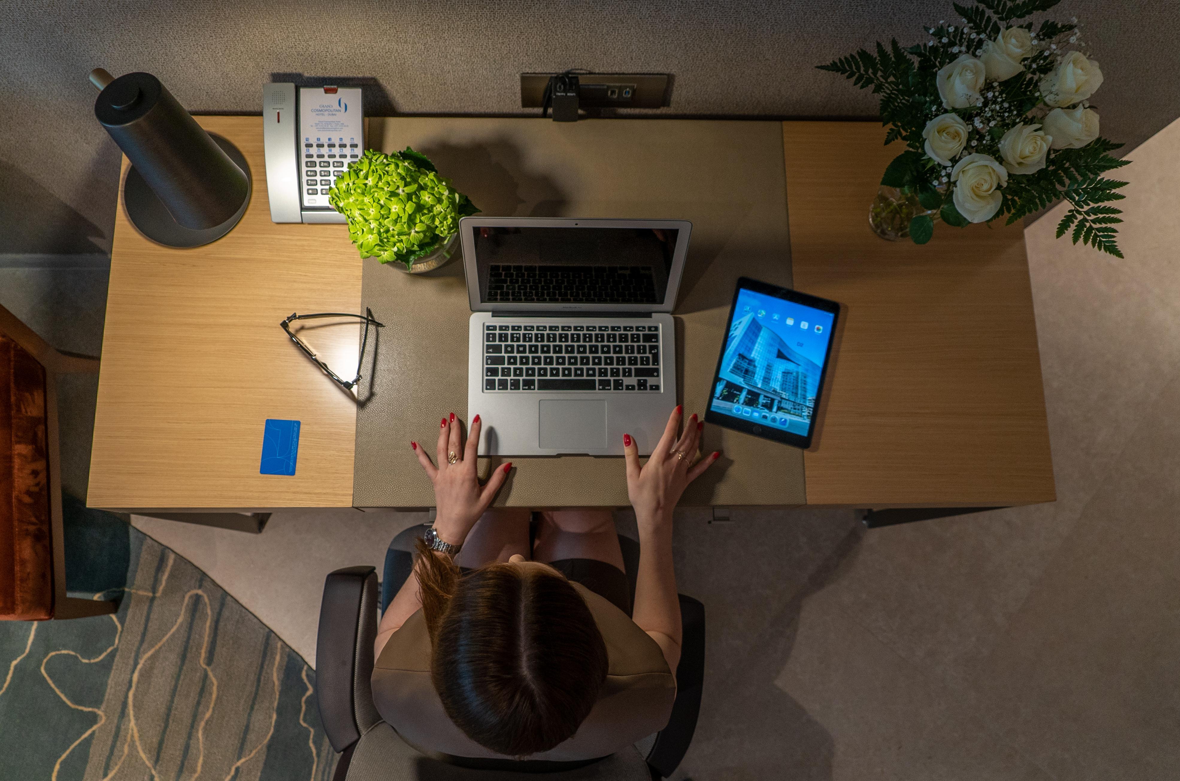 迪拜大都会大酒店 外观 照片 The photo shows a top-down view of a modern workspace. A person with long hair is sitting at a wooden desk, using a laptop with an open screen. Next to the laptop, there is a tablet displaying a cityscape. The desk also features a green potted plant,