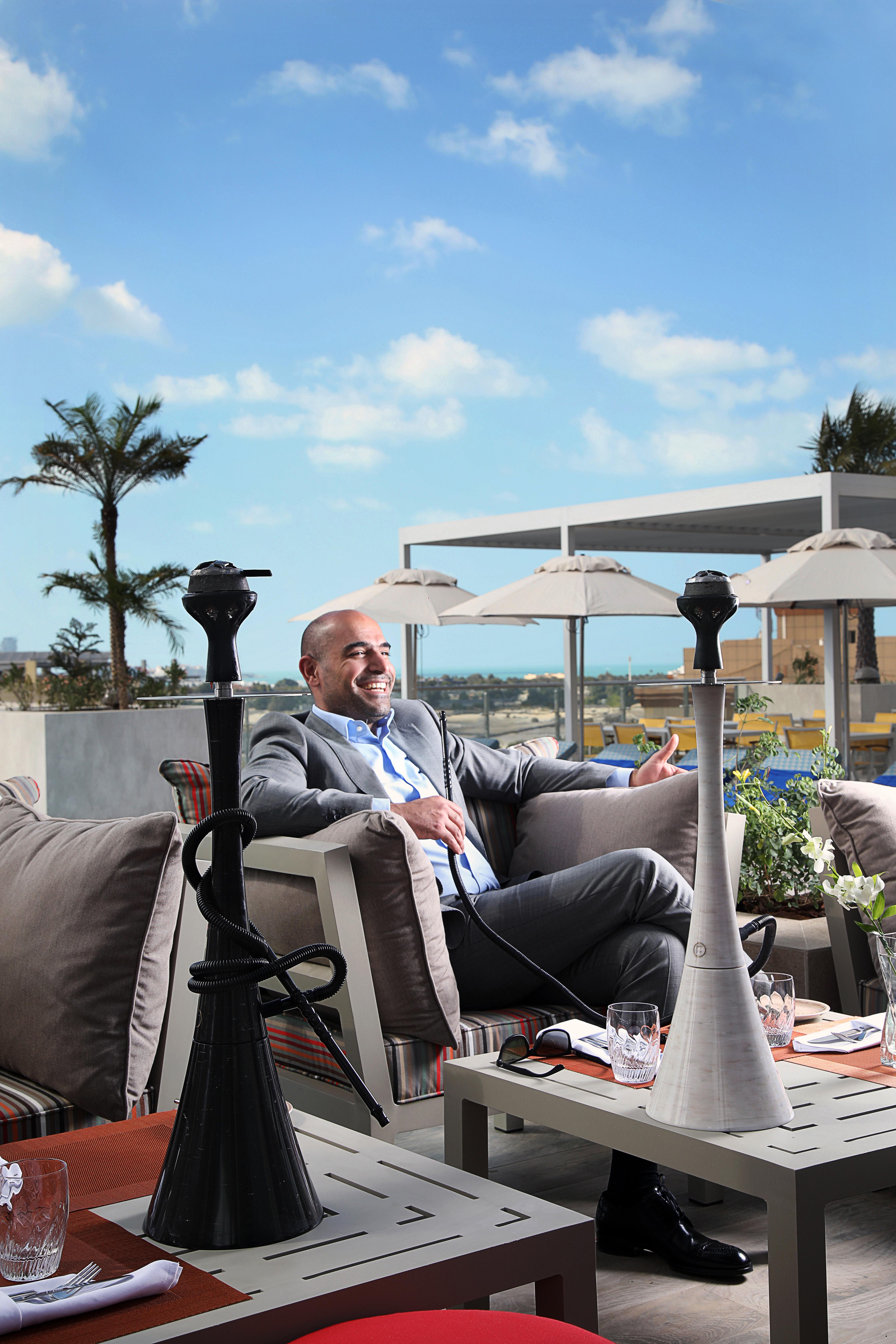 迪拜大都会大酒店 外观 照片 The image shows a man sitting comfortably on a sofa in an outdoor lounge area. He is dressed in a suit and is smiling, suggesting a relaxed atmosphere. In front of him are two hookahs, which are traditional smoking devices. The background features pa