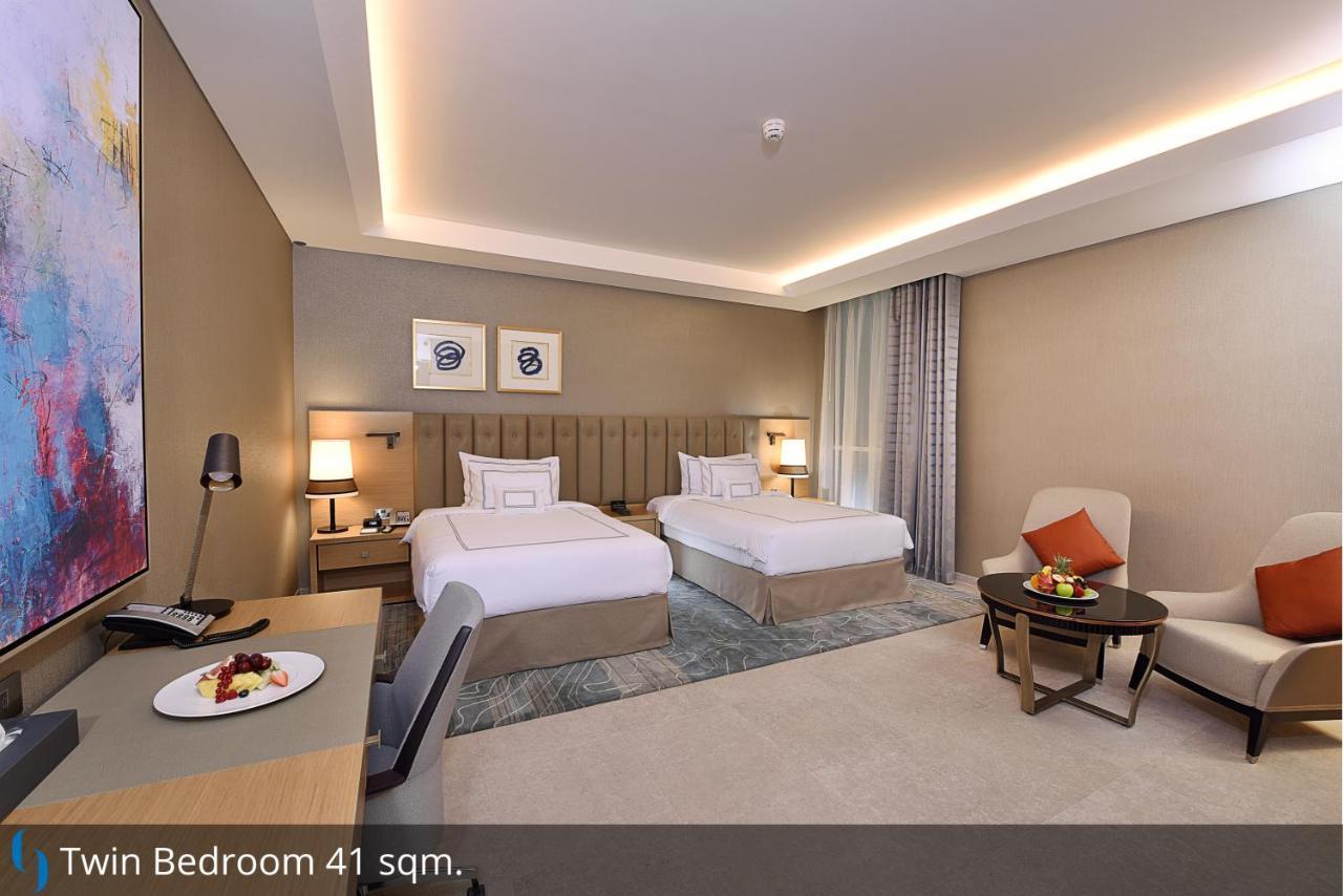 迪拜大都会大酒店 外观 照片 The photo shows a modern twin bedroom with two beds. The beds are neatly made, and there’s a light-colored rug on the floor. In the corner, there's a small table with a chair, and on the table, there's a plate of fruit. The decor features a neutral c
