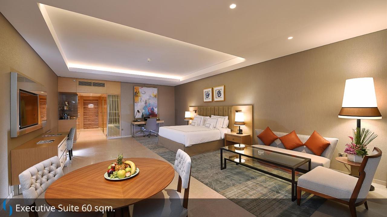 迪拜大都会大酒店 外观 照片 The image shows a spacious hotel suite. It features a king-sized bed with a modern design, accompanied by decorative pillows. To one side, there is a seating area with a sofa and a glass coffee table. A dining table with chairs is also visible, posit