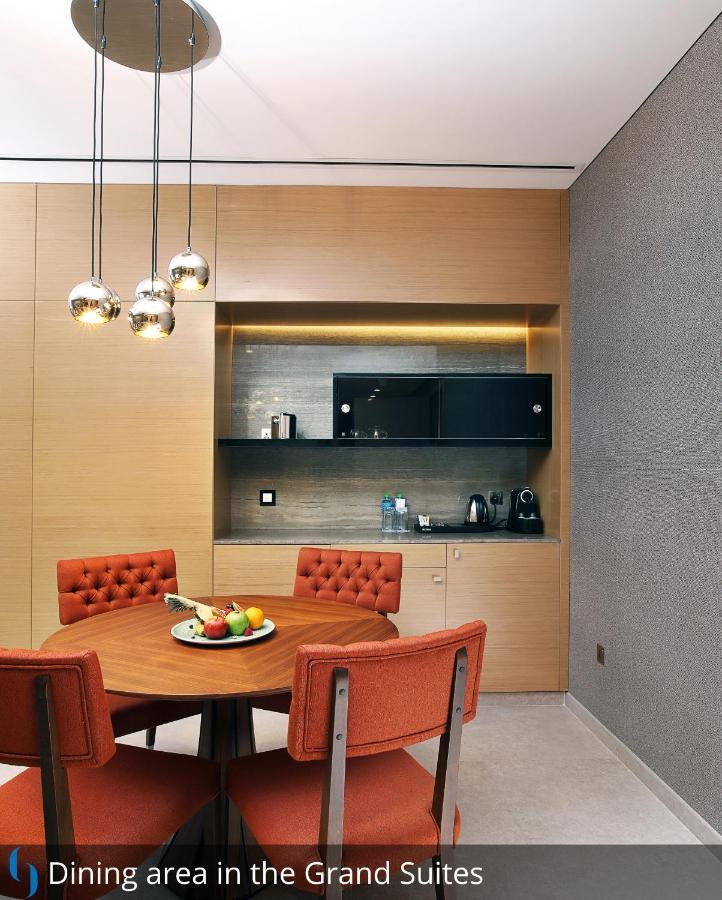 迪拜大都会大酒店 外观 照片 The photo depicts a modern kitchen area. There is a round dining table with orange upholstered chairs surrounding it. On the table, a bowl of assorted fruits is displayed. In the background, a countertop with some kitchen appliances, including a kett