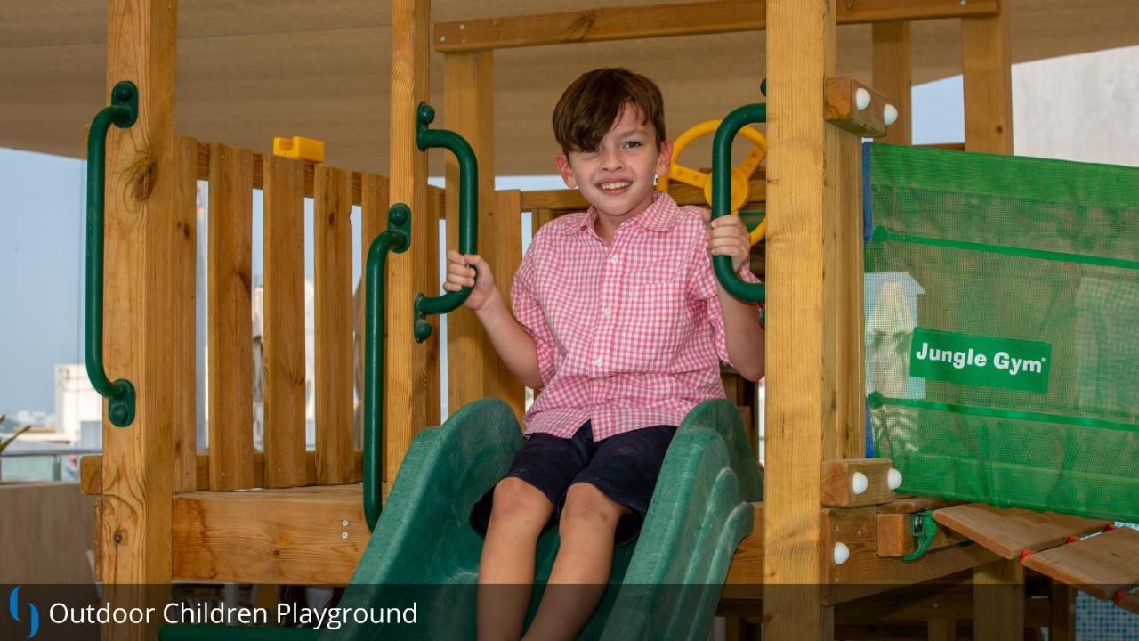 迪拜大都会大酒店 外观 照片 The image shows a young boy sitting on a slide in a children's playground. He is wearing a pink checkered shirt and dark shorts. The playground equipment is made of wood and features green sliding elements. The boy has a joyful expression, suggesting