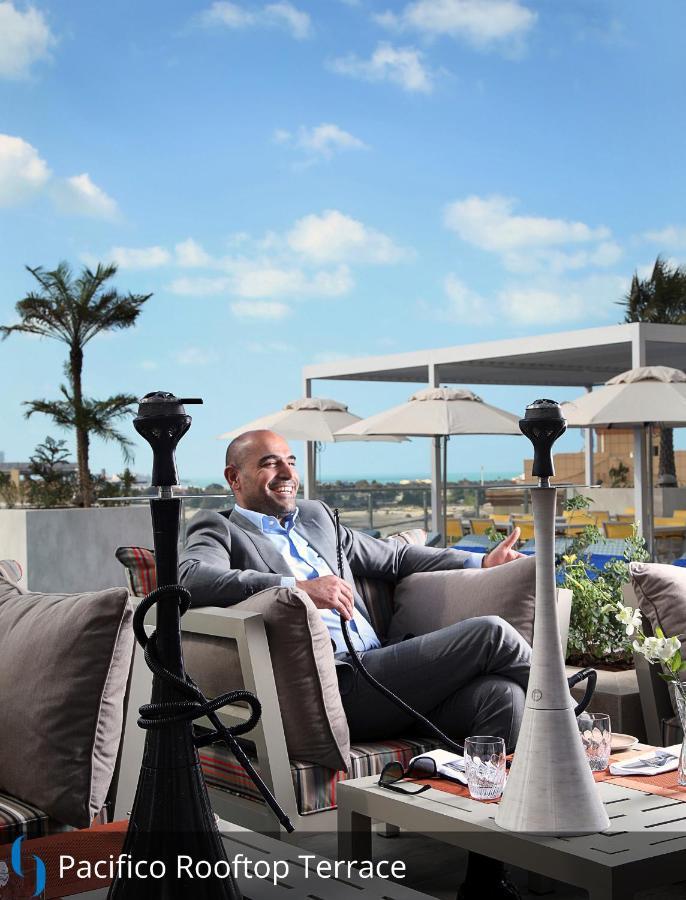 迪拜大都会大酒店 外观 照片 The photo shows a man sitting comfortably in an outdoor lounge area. He is dressed in a light grey suit and appears to be smiling and relaxed. In the background, there are several palm trees and sun loungers with umbrellas, suggesting a tropical or u