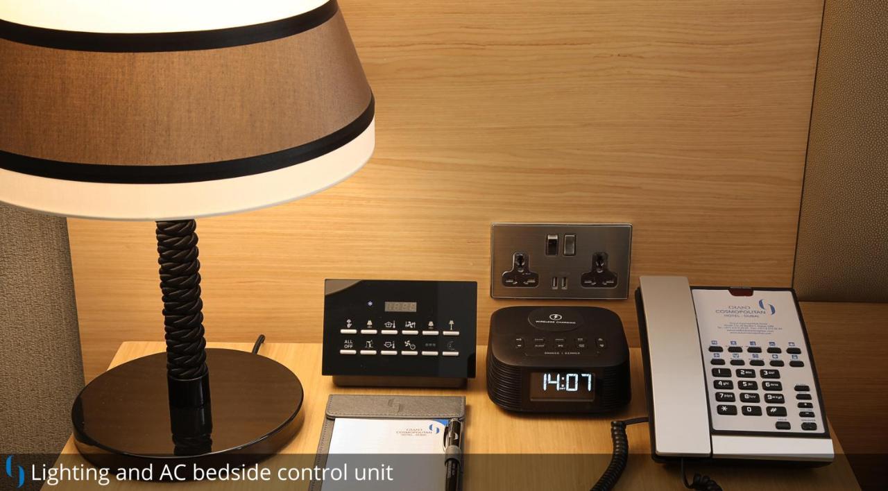 迪拜大都会大酒店 外观 照片 The photo shows a bedside setup with several items. There is a table lamp with a dark lampshade on the left. Next to it is a control unit with various buttons, likely for managing room settings such as lighting and air conditioning. Beside the contro