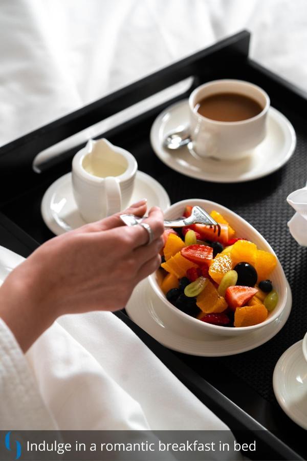 迪拜大都会大酒店 外观 照片 The photo displays a hand holding a fork, poised to pick up fruit from a bowl. The bowl contains a colorful assortment of fresh fruit, including pieces of orange, strawberries, grapes, and blueberries. Nearby, there is a cup of coffee and a small sau