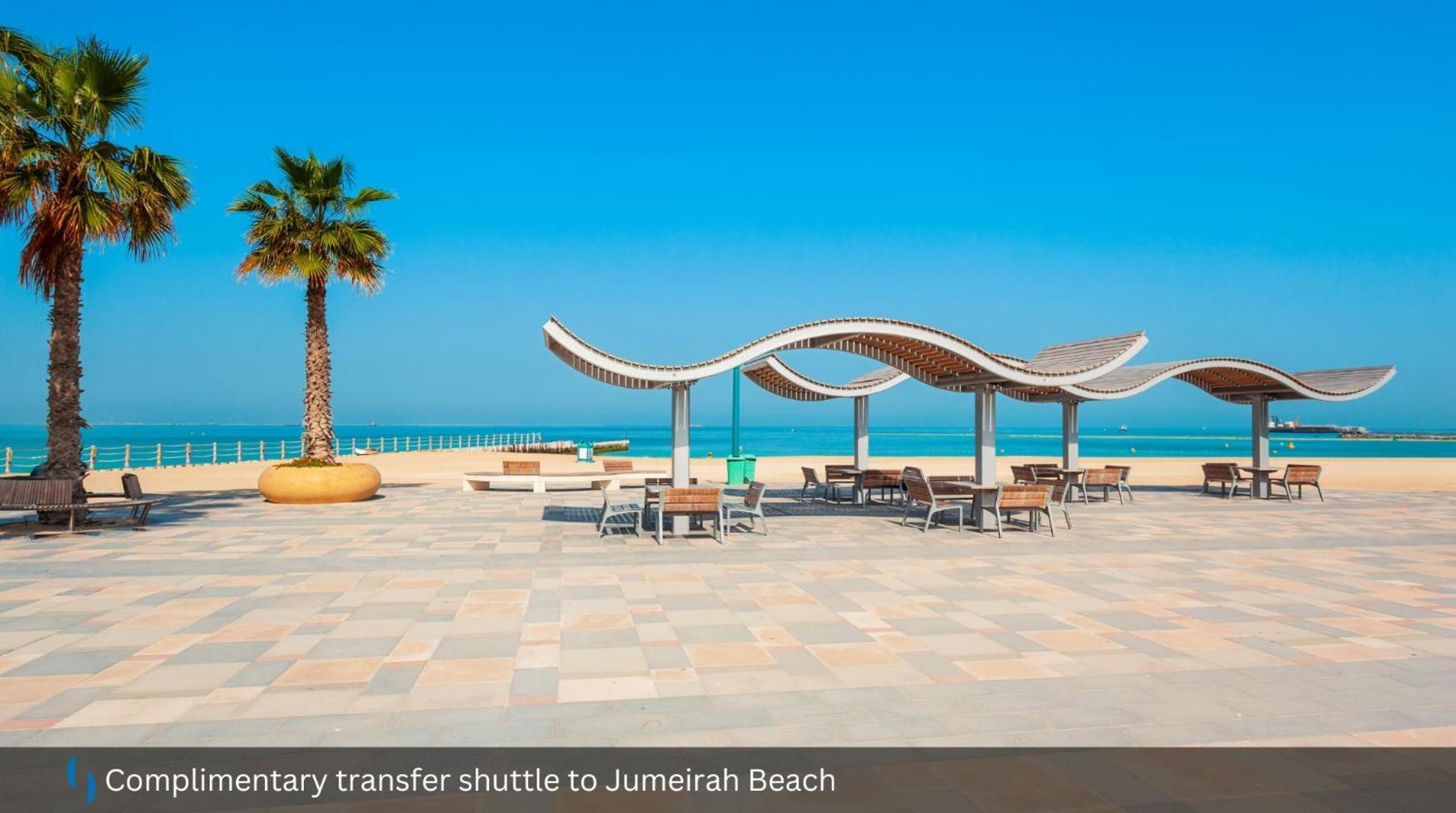 迪拜大都会大酒店 外观 照片 The photo shows a scenic beach area, likely at Jumeirah Beach. In the foreground, there are wooden benches and tables arranged under shaded structures with a wavy design. A tall palm tree adds to the tropical feel of the setting. In the background, y