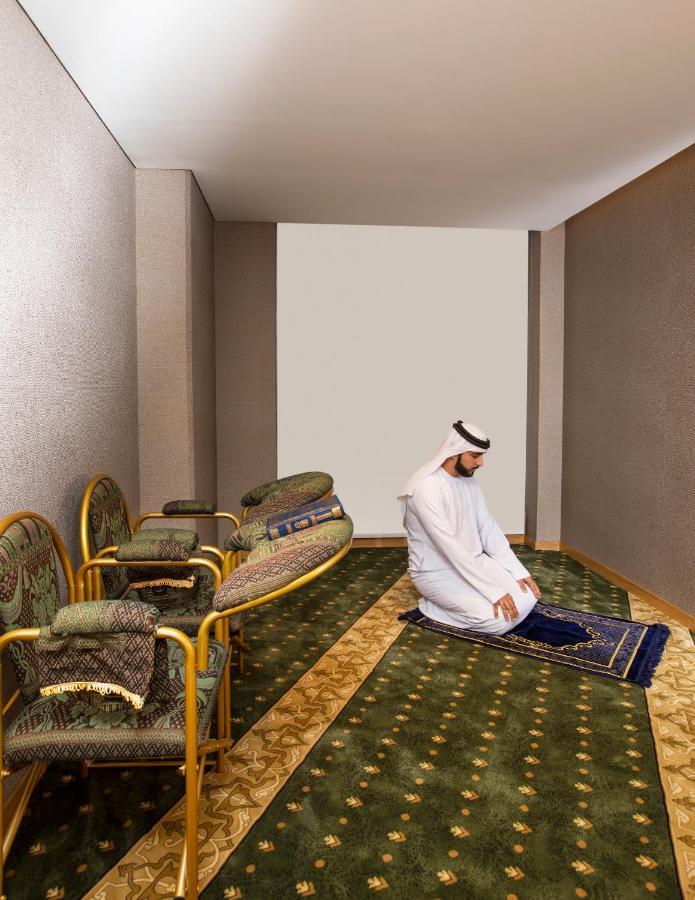 迪拜大都会大酒店 外观 照片 The image shows a simple, serene indoor setting. In the center, a man wearing traditional attire is kneeling on a prayer rug, engaged in prayer. Surrounding him are several ornate chairs, and the floor is adorned with patterned carpets. The walls are